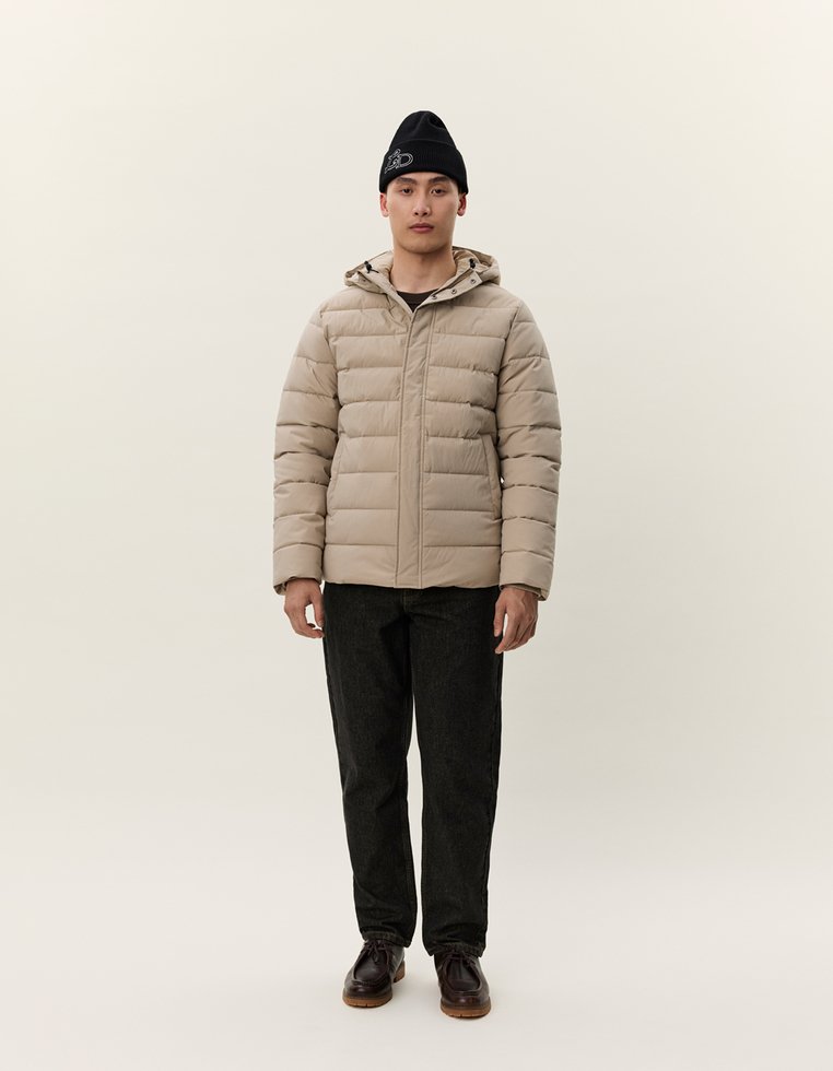 Mann iført beige puffjakke, svart beanie, svarte bukser og mørkebrune sko som står foran en ensfarget lys bakgrunn.