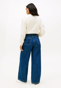 Niebieskie dżinsy o szerokich nogawkach, z wysokim stanem i przeszytymi tylnymi kieszeniami, zestawione z kremowym swetrem, widziane od tyłu na jasnym tle.