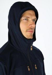 Marineblå softshell-jakke med høj krave og hætte, der har to lynlåslommer med brune detaljer og en glat, struktureret overflade.