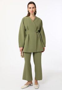 Olive green two-piece outfit featuring a wrap-style top with wide sleeves, belted waist, and flared pants. Includes cream-colored mules.