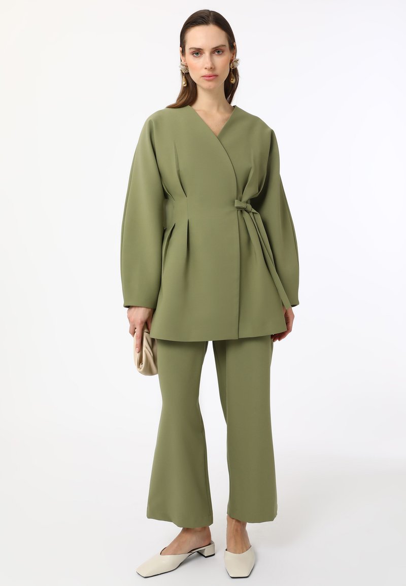 Olijfgroene twee-delige outfit met een wikkelstijl top met brede mouwen, een ceintuur in de taille en uitlopende broekspijpen. Inclusief crèmekleurige muiltjes.