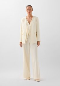 Traje crema que incluye un blazer estructurado con dos bolsillos y pantalones de pierna ancha, hecho de una tela texturizada, combinado con zapatos a juego.