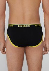 Ropa interior para hombres en negro con acentos amarillos, con una cinturilla ancha que presenta la marca "TEZENIS" y un diseño ajustado con aberturas en las piernas curvadas.