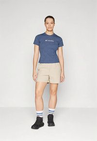 Kvinne som står iført en tettsittende blå Columbia t-skjorte, beige shorts, hvite sokker med blå striper og svarte vandringssko mot en enkel bakgrunn.