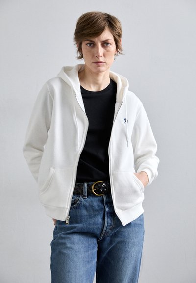 Un sweat à capuche zippé blanc avec des poches avant, porté sur un t-shirt noir, associé à un jean bleu et une ceinture noire tressée. Tissu texturé.