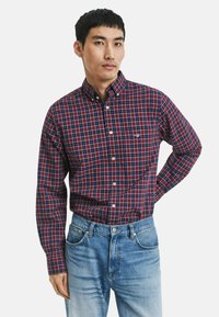 Mann in einem rot-blauen karierten Hemd, das in eine hellblaue Jeans gesteckt ist, steht mit einer Hand hinter seinem Rücken.