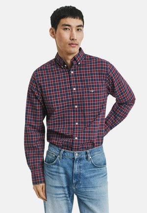 Mann in einem rot-blauen karierten Hemd, das in eine hellblaue Jeans gesteckt ist, steht mit einer Hand hinter seinem Rücken.