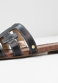 Schwarze Ledersandale mit ausgeschnittenem Design, weißer Naht und goldenem Logoakzent am Riemen. Strukturierter brauner Fußbett.