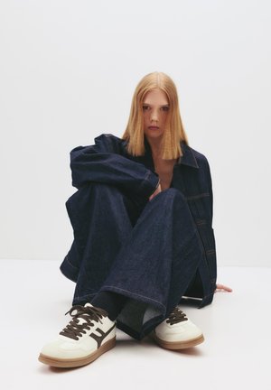 Junge Frau mit glatten blonden Haaren sitzt im Schneidersitz und trägt eine dunkle Jeansjacke und Hose mit weißen Turnschuhen.