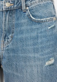 Jean en denim bleu avec bouton métal, poche avant, et petites déchirures usées près de la poche et de la cuisse.