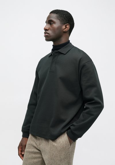 Άνδρας που φοράει ένα μαύρο μακρυμάνικο πόλο μπλουζάκι πάνω από έναν μαύρο τερτλένκ και μπεζ υφασμάτινο παντελόνι, στέκεται με το ένα χέρι στην τσέπη.