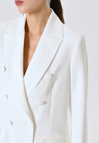 Witte dubbelzijdige blazer met een diepe V-hals, textuurstof en grote met stof bedekte knopen. Getailleerde pasvorm en strakke lijnen.