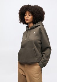 Olivovo zelená mikina s kapucňou na šnúrky, predným vreckom a malým logom. Mäkká textúra a voľný strih, doplnená o béžové nohavice.