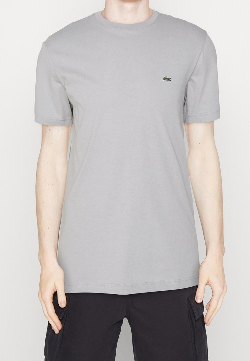 Homme portant un T-shirt à manches courtes uni gris clair avec un petit logo de crocodile vert sur la poitrine gauche, associé à un short cargo noir.