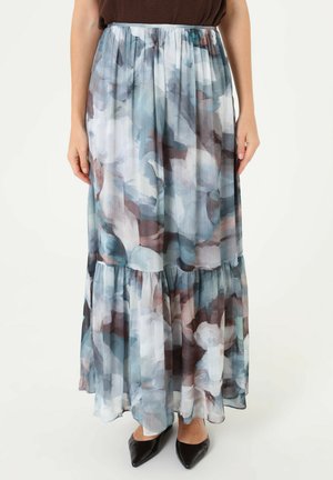 Femme portant une longue jupe fluide avec des motifs floraux abstraits bleus, gris et marron et des chaussures noires à bout pointu.