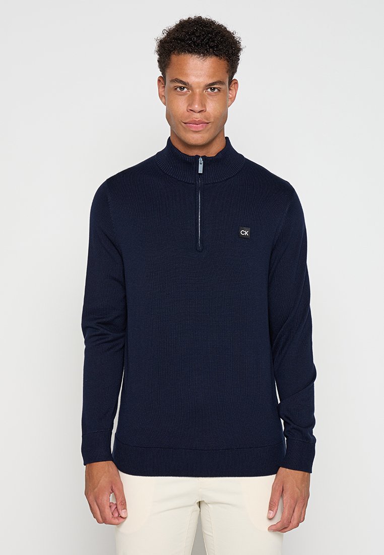 Pull-over zippé bleu marine avec une texture côtelée et un col haut ; comporte un petit logo CK sur la poitrine ; design ajusté.