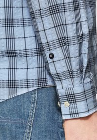 Lichtblauwe geruite shirt met zwart en grijs plaidpatroon. Voorzien van knoopsluitingen op de manchetten en een gestructureerde stof. Gecombineerd met jeans.