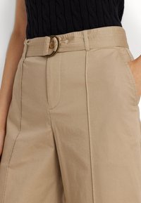 Beige trousers with a wide-leg cut, featuring a belt with a round metal buckle, side pockets, and a smooth fabric texture.