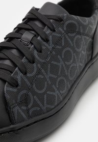 Baskets noires avec une tige en tissu texturé présentant un motif de logo subtil et des lacets plats noirs. Semelle extérieure en caoutchouc avec une finition lisse.