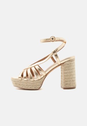 Zlaté sandále na platforme s remienkami, prackou na členku a hrubým pleteným jútovým blokovým podpätkom, z boku na bielom pozadí.