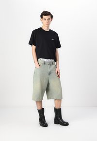 Schwarzes T-Shirt mit kurzen Ärmeln und Logo, hellblau gewaschene, weite Denim-Shorts und schwarze Stiefeletten mit Schnallen. Neutraler Hintergrund.