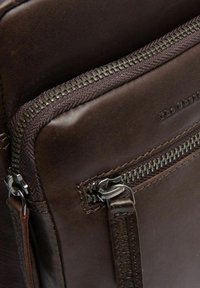 Sac en cuir marron avec une fermeture éclair en métal visible, finition texturée et forme plate. La marque est embossée sur la surface en cuir.