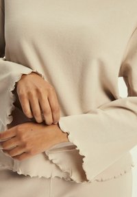 Beżowa bluzka z dzianiny wafelowej, z rozszerzanymi rękawami i falbanowymi mankietami, prezentująca teksturowaną powierzchnię oraz miękki, luźny krój.