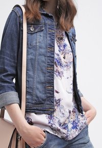 Jeansjacka med två bröstfickor, slitna detaljer och uppvikta ärmar, över en blus med blommönster. Ljus färgad handväska synlig.