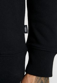 Sudadera negra con puños acanalados, una franja blanca de acento en el costado y una etiqueta del logo de Vans visible en la manga.