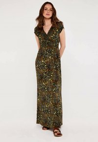 Maxikleid aus grünem Stoff mit einem gemischten Muster aus Blättern und Tierdruck, V-Ausschnitt und kurzen Ärmeln. Getragen mit braunen Sandalen.