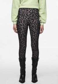 Legíny s tmavým leopardím potiskem v kombinaci s černými kovbojskými botami a světle zeleným dlouhým rukávem na bílém pozadí.