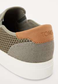 Pelēkā sieta kedas ar ādas papēža akcentu, uz kura ir uzraksts "TOMS". Izceļas ar teksturētu virsmu un baltu gumijas zoli.