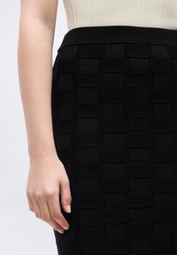 Κοντινό ενός χεριού ατόμου που ακουμπάει στο πλάι, φοράει μπεζ μπλούζα και μαύρη υφασμάτινη φούστα με υφαντό μοτίβο.