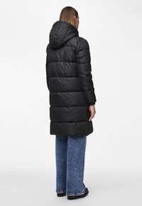 Cappotto imbottito nero con cappuccio, caratterizzato da un design trapuntato, lunghezza lunga e finitura testurizzata. Abbinato a jeans blu a gamba larga.