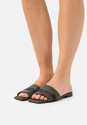 Pieds nus dans des sandales à glissière vert foncé avec des sangles noires et des semelles carrées sur un fond blanc.