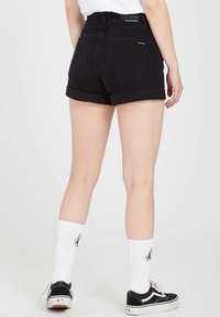Shorts en denim noir taille haute avec ourlets retroussés, dotés de poches arrière et du logo Volcom. Portés avec des chaussettes blanches et des baskets noires.