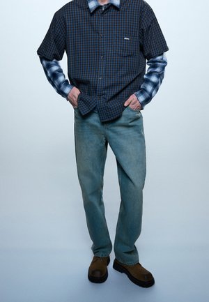 Personne portant des chemises à carreaux bleues superposées, un jean bleu clair et des chaussures marron, debout les mains dans les poches devant un fond uni.