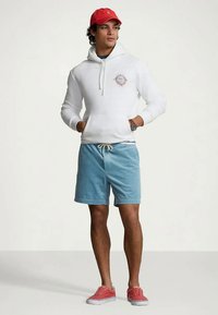 Jovem numa hoodie branca, calções azuis, boné vermelho e tênis vermelhos de pé com as mãos nos bolsos contra um fundo simples.