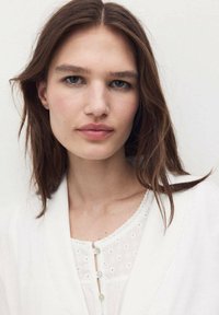 Bluză albă cu cusături decorative și nasturi, având un material texturat dedesubt. Păr castaniu și ten curat cu machiaj neutru.