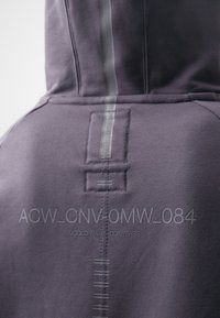 Gros plan de l'arrière d'un sweat à capuche violet montrant une couture réfléchissante verticale et le texte « ACW_CNV-0MW_084 » et « A-COLD-WALL* CONVERSE ».