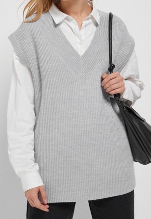 Personne portant une chemise blanche à col sous un pull sans manches en maille côtelée gris clair, tenant un sac à bandoulière en cuir noir avec une sangle nouée.