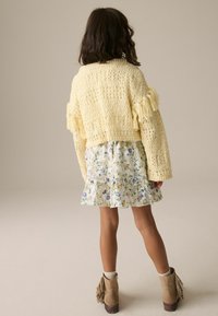 Gelbe gestrickte Cardigan mit Fransen, kombiniert mit einem floralen Minikleid und beigen Stiefeletten. Der Hintergrund ist neutral.