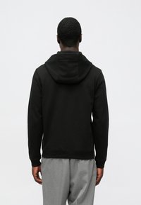 Camisola preta com capuz, bolso canguru, capuz com cordão e punhos canelados. Textura de tecido lisa, usada com calças cinzentas plissadas.