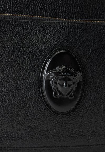 Surface en cuir noir texturé avec un emblème ovale noir présentant un design en relief de tête de Méduse et une fermeture éclair dorée sur le bord supérieur.