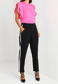 Camiseta corta de manga corta en un brillante color rosa, con detalles de volantes, combinada con pantalones negros ajustados que tienen líneas blancas a los lados y zapatos de tacón negro de punta.