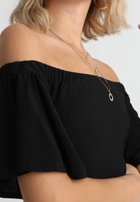 Haut noir à épaules dénudées avec manches courtes et évasées. Tissu lisse, coupe décontractée, agrémenté de colliers superposés et de boucles d'oreilles circulaires.