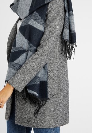 Personne portant un manteau gris texturé et une grande écharpe à carreaux bleus et gris avec des franges, associée à un jean bleu.