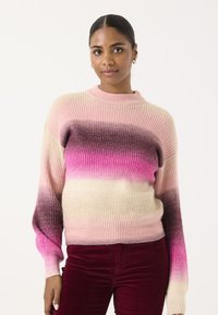 Strickpullover mit einem Farbverlauf von hellem Rosa zu Lila, mit gerippter Textur und hohem Kragen. Das Modell trägt dazu bordeauxfarbene Hosen.