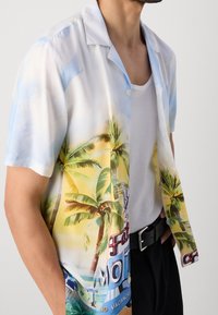 Camisa de manga corta en tejido liviano y brillante con un estampado vibrante de palmeras y atardecer en tonos amarillos y azules. Botones blancos visibles.