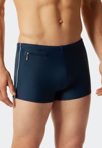 Marinefarbene Badehorts aus elastischem Material, mit einer kleinen Reißverschlusstasche und einem weißen Streifen an der Seite, enger Schnitt und glatter Textur.